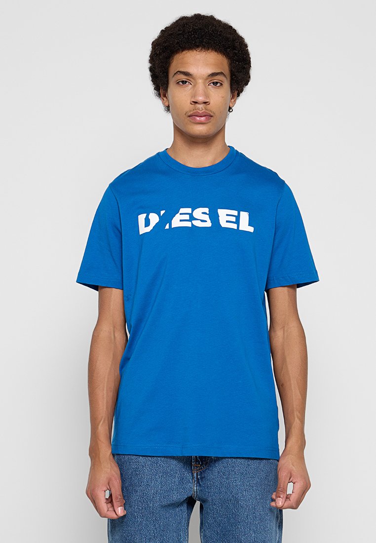 Junger Mann mit lockigem Haar, der ein leuchtend blaues Diesel-T-Shirt und blaue Jeans trägt, vor einem schlichten hellgrauen Hintergrund steht.