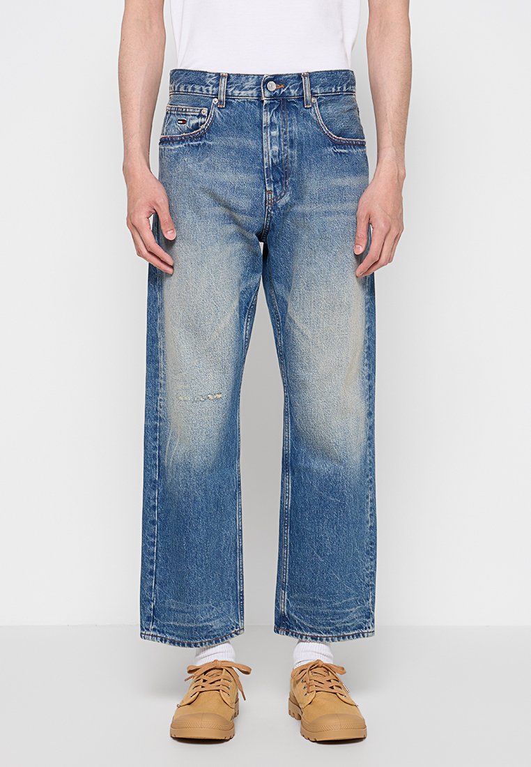 Person, die weit geschnittene, blaue verwaschene Jeans mit leichten Abnutzungen, weiße Socken, kastanienbraune Schnürschuhe und ein in die Hose gestecktes weißes Hemd trägt.