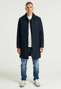 Marineblauer langer Mantel mit Knopfverschluss, seitlichen Taschen und Kragen. Getragen über einem weißen Hemd und kombiniert mit hellblauen Jeans und Turnschuhen.