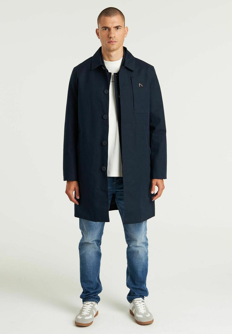 Marineblauer langer Mantel mit Knopfverschluss, seitlichen Taschen und Kragen. Getragen über einem weißen Hemd und kombiniert mit hellblauen Jeans und Turnschuhen.