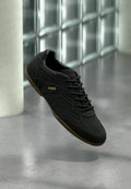 Schwarzer Sneaker mit niedrigem Schaft, brauner Sohle, schwarzen Schnürsenkeln und "HUGO"-Logo an der Seite, präsentiert vor einem Hintergrund aus Glasbausteinen.