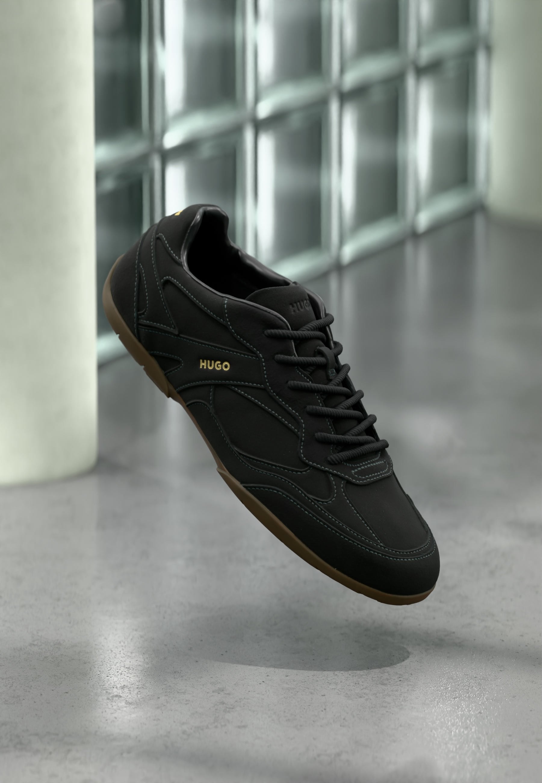 Schwarzer Sneaker mit niedrigem Schaft, brauner Sohle, schwarzen Schnürsenkeln und "HUGO"-Logo an der Seite, präsentiert vor einem Hintergrund aus Glasbausteinen.