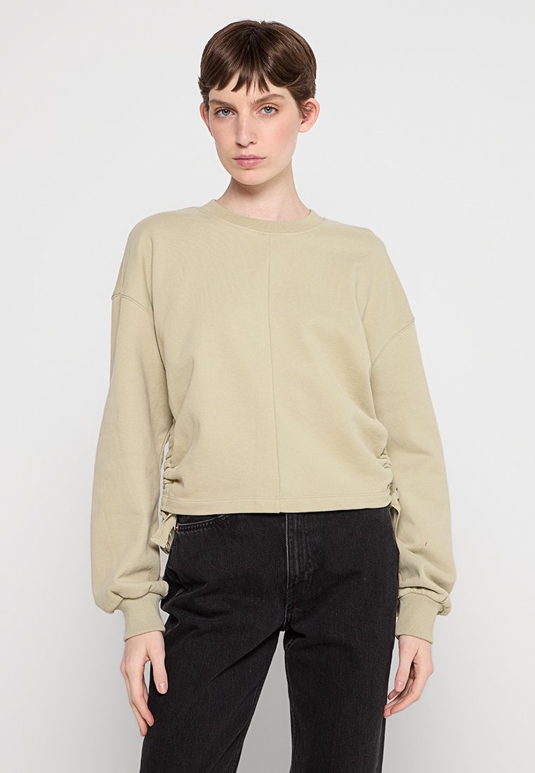 Beige verkürzter Pullover mit langen Ärmeln, gerippten Bündchen, seitlichen Raffungselementen und rundem Ausschnitt. Getragen mit schwarzen Hosen.