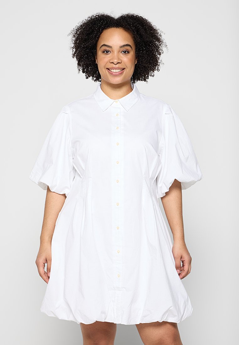 Lächelnde Frau mit lockigem Haar, die ein weißes Hemdkleid mit Puffärmeln und einem gewellten Saum trägt.