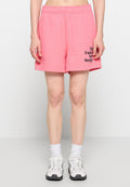Person trägt pinke Shorts mit schwarzem Text am linken Bein, weiße Socken und weiße Laufschuhe, steht vor einem einfarbigen hellen Hintergrund.