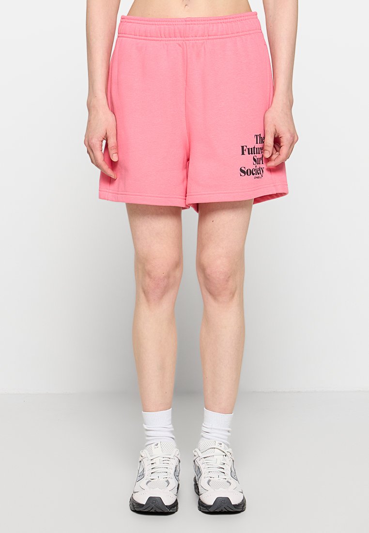 Person trägt pinke Shorts mit schwarzem Text am linken Bein, weiße Socken und weiße Laufschuhe, steht vor einem einfarbigen hellen Hintergrund.