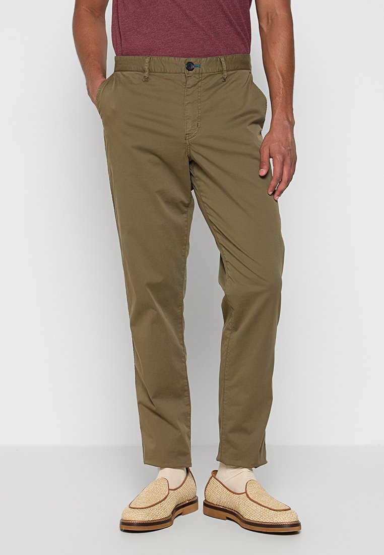 Ein Mann trägt ein kastanienrotes T-Shirt und olivgrüne Hosen, steht selbstbewusst mit einer Hand in seiner Tasche und trägt hellfarbene Slipper.