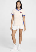 Beige Nike-Trikot mit blauen und roten Akzenten sowie einem V-Ausschnitt, kombiniert mit einem weißen Plisseerock und weißen Sneakers.