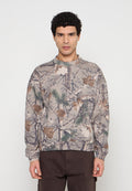 Mann trägt ein langärmeliges, beiges Camouflage-Sweatshirt mit Baumzweigen, Blättern und Kiefernnadeln und steht vor einem schlichten Hintergrund.