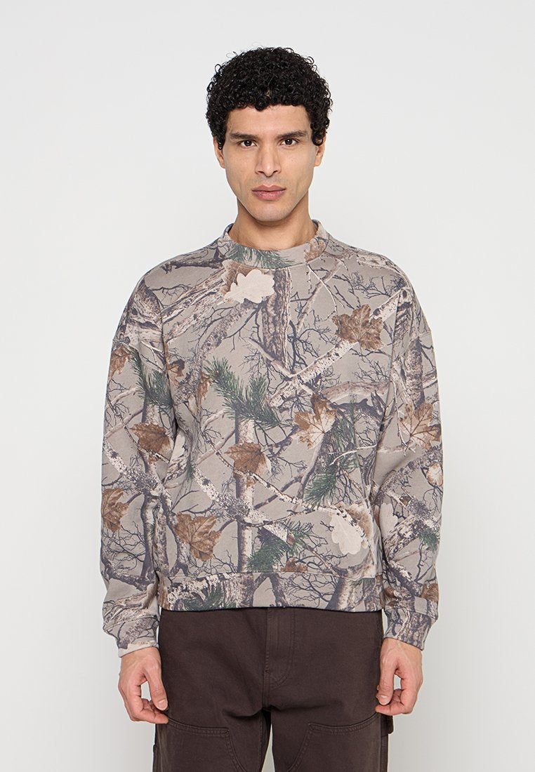Mann trägt ein langärmeliges, beiges Camouflage-Sweatshirt mit Baumzweigen, Blättern und Kiefernnadeln und steht vor einem schlichten Hintergrund.
