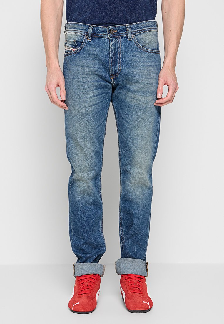 Mann trägt ausgewaschene blaue Jeans, die an den Säumen aufgerollt sind, und rote Puma-Sneaker, steht vor einem schlichten hellgrauen Hintergrund.