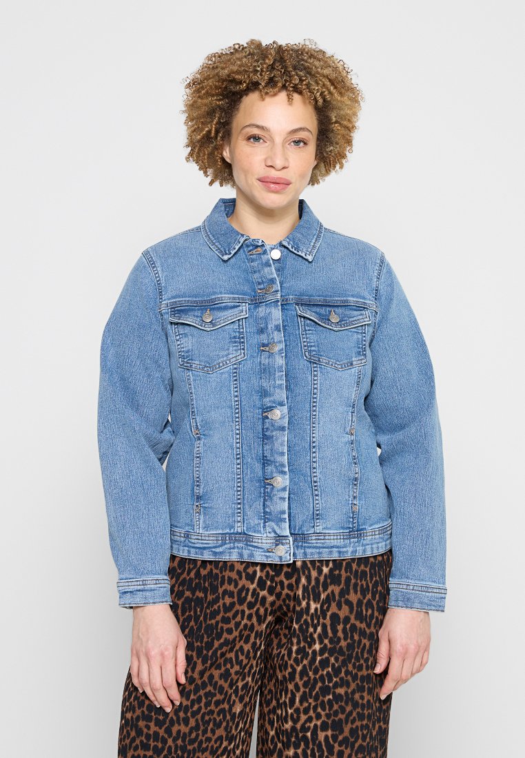 Eine Person trägt eine hellblaue Jeansjacke mit Knopfleiste und Leopardenmuster-Hose, steht vor einem einfarbigen hellen Hintergrund.