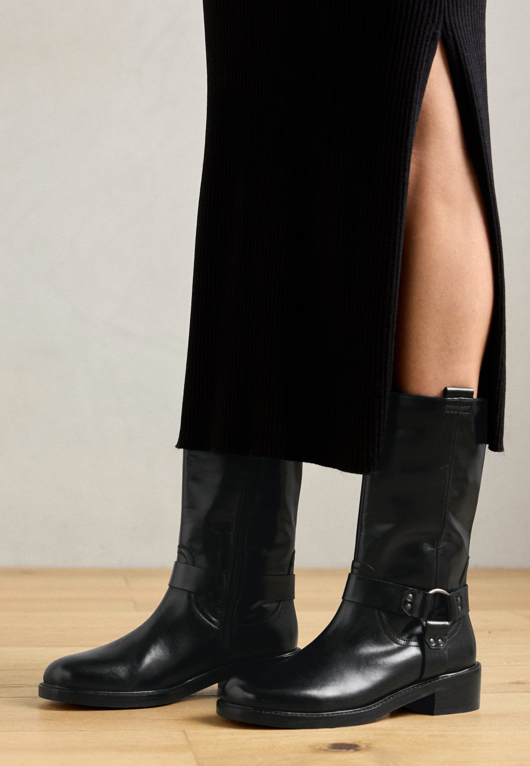 Schwarze Leder-Stiefel bis zum Knie mit runder Spitze, glatter Oberfläche, flachem Absatz, seitlichem Reißverschluss und einem verstellbaren Riemen mit Schnallen-Detail.