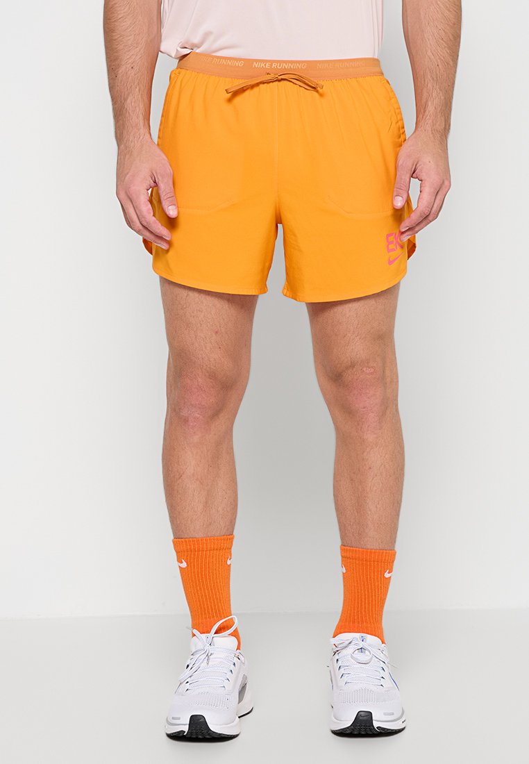 Mann trägt leuchtend orangefarbene Nike-Laufshorts, passende orange Nike-Socken und weiße Laufschuhe, steht vor einem schlichten Hintergrund.
