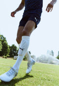 Weiße Fußballschuhe mit grauen Akzenten, marineblaue Sportshorts und weiße knielange Socken mit einem Logo, vor einem Rasenfeld.