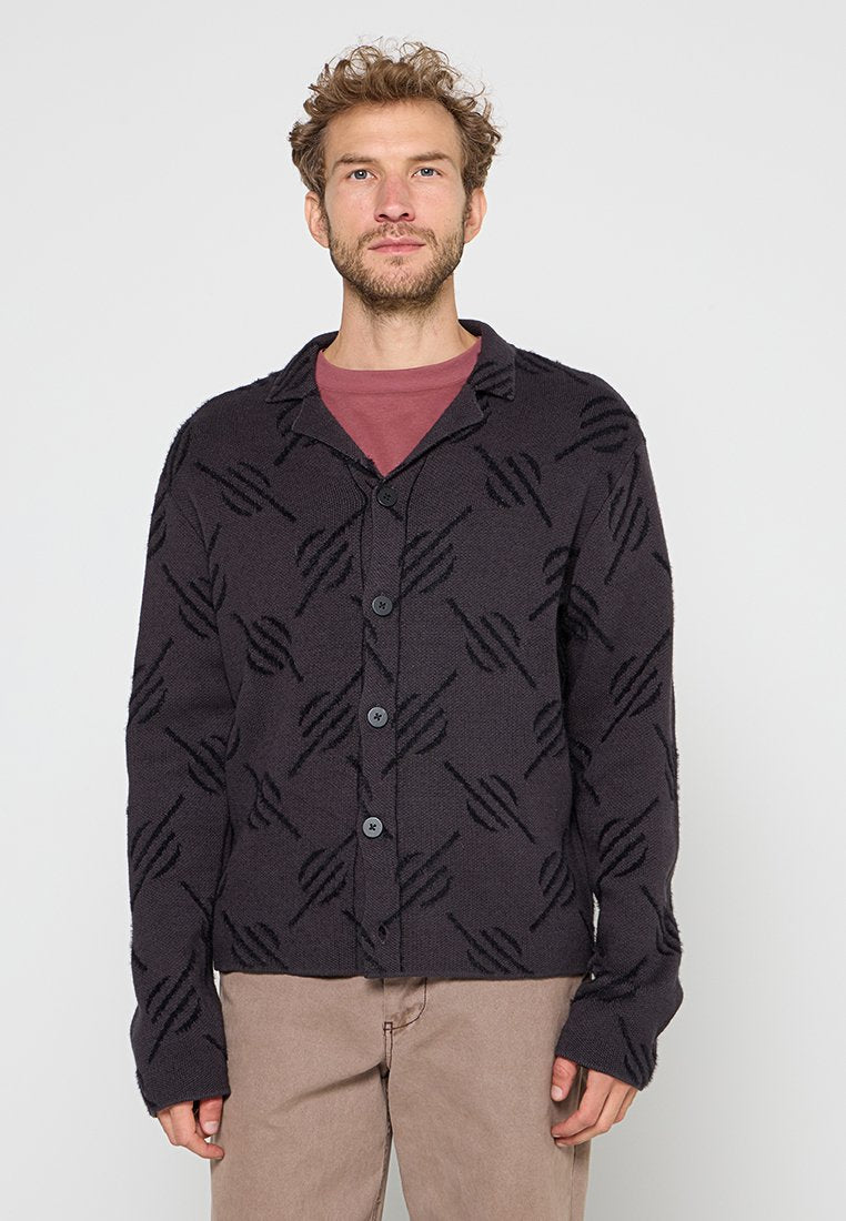 Mann mit lockigem Haar, der einen schwarzen, geknöpften Cardigan mit einem gezackten Muster über einem kastanienbraunen Hemd und beigen Hosen trägt, steht vor einem weißen Hintergrund.