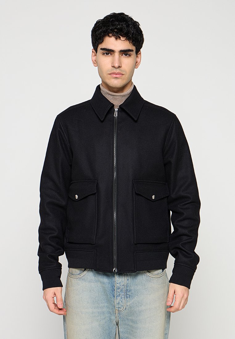 Schwarze Reißverschlussjacke aus strukturiertem Stoff mit Kragen, zwei Fronttaschen und figurbetontem Schnitt. Über einem Rollkragenpullover getragen.