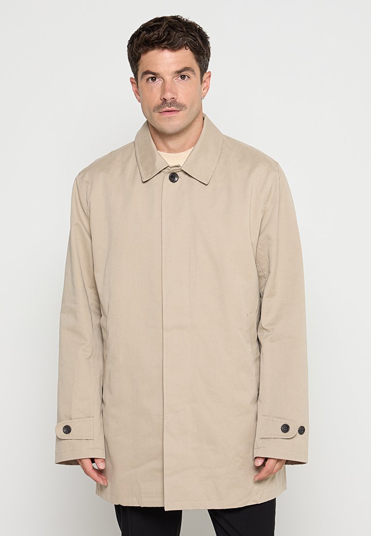 Mann in beige Knopfjacke mit Kragen, der vor einem schlichten weißen Hintergrund steht.