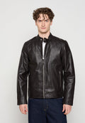 Junger Mann mit lockigem Haar, der eine schwarze Lederjacke mit Reißverschluss und dunkelblaue Jeans trägt, steht vor einem einfarbigen hellen Hintergrund.