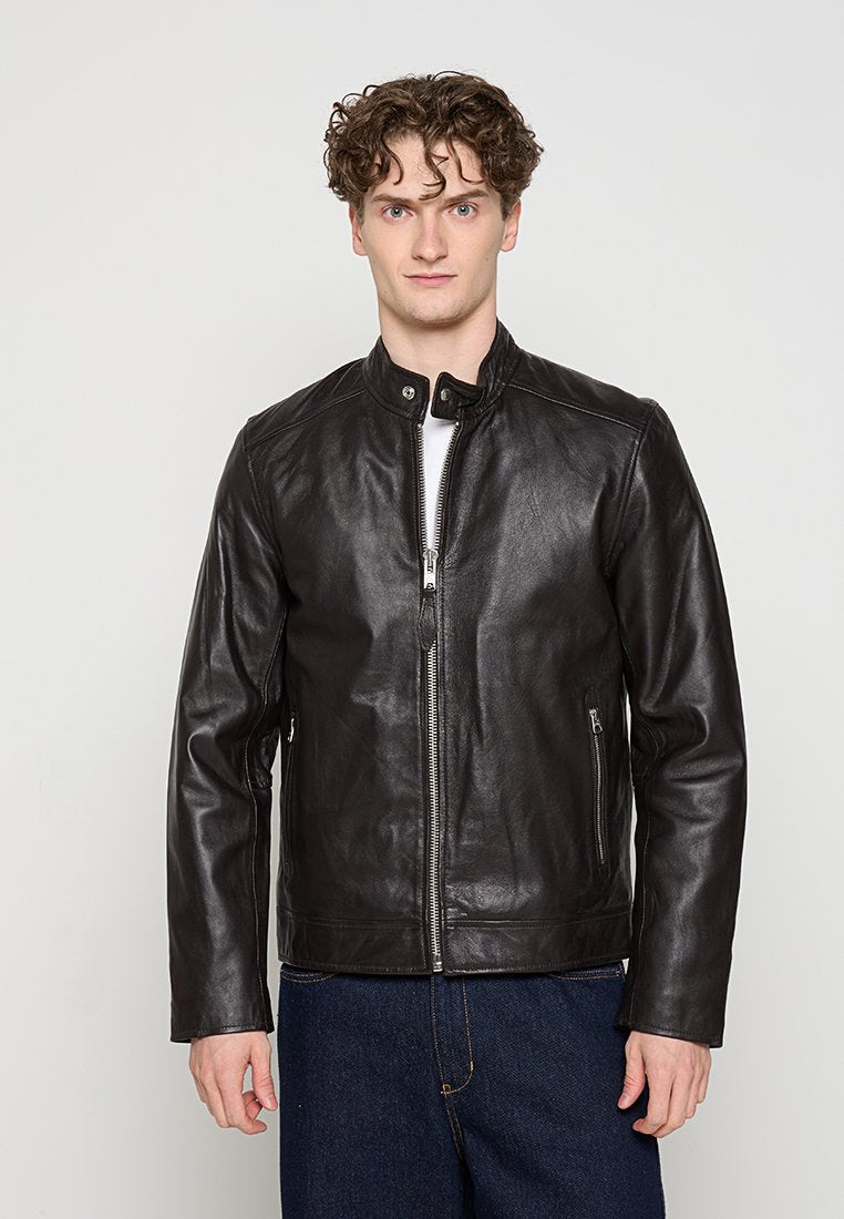 Junger Mann mit lockigem Haar, der eine schwarze Lederjacke mit Reißverschluss und dunkelblaue Jeans trägt, steht vor einem einfarbigen hellen Hintergrund.