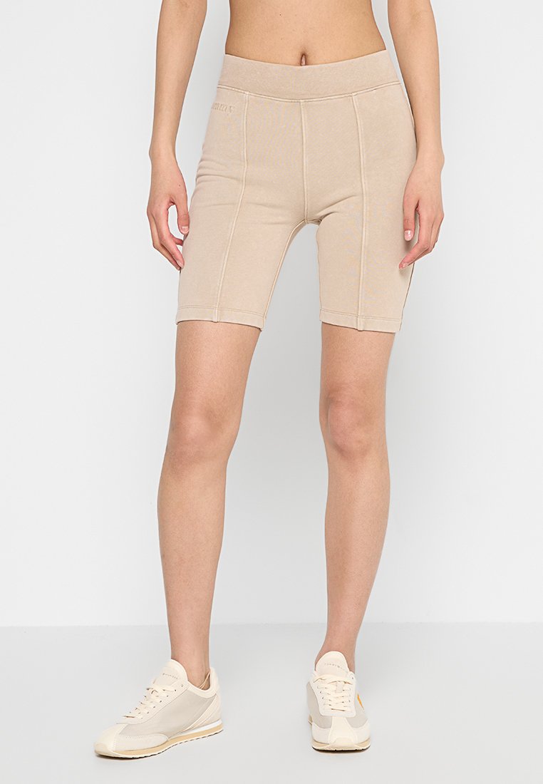 Beige Radshorts mit hohem Taillenbund, elastischem Baumwollmaterial, kontrastierenden Nähten und figurbetontem Schnitt, kombiniert mit hellen Sneakers.