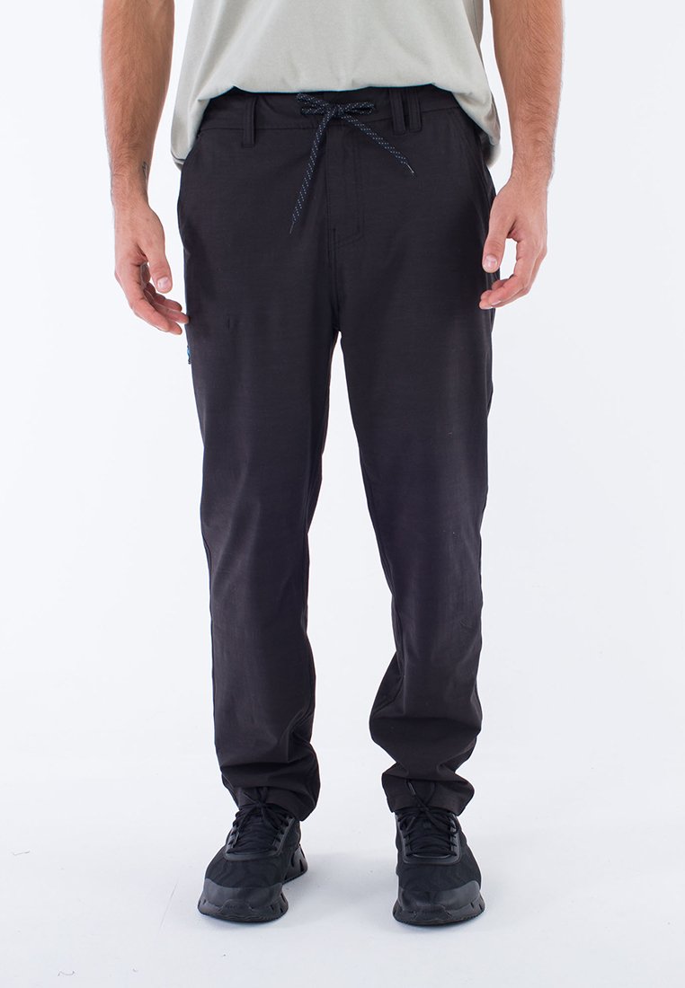 Mann trägt schwarze lässige Hose mit Kordelzugbund und schwarze Sneakers, steht vor einem weißen Hintergrund.