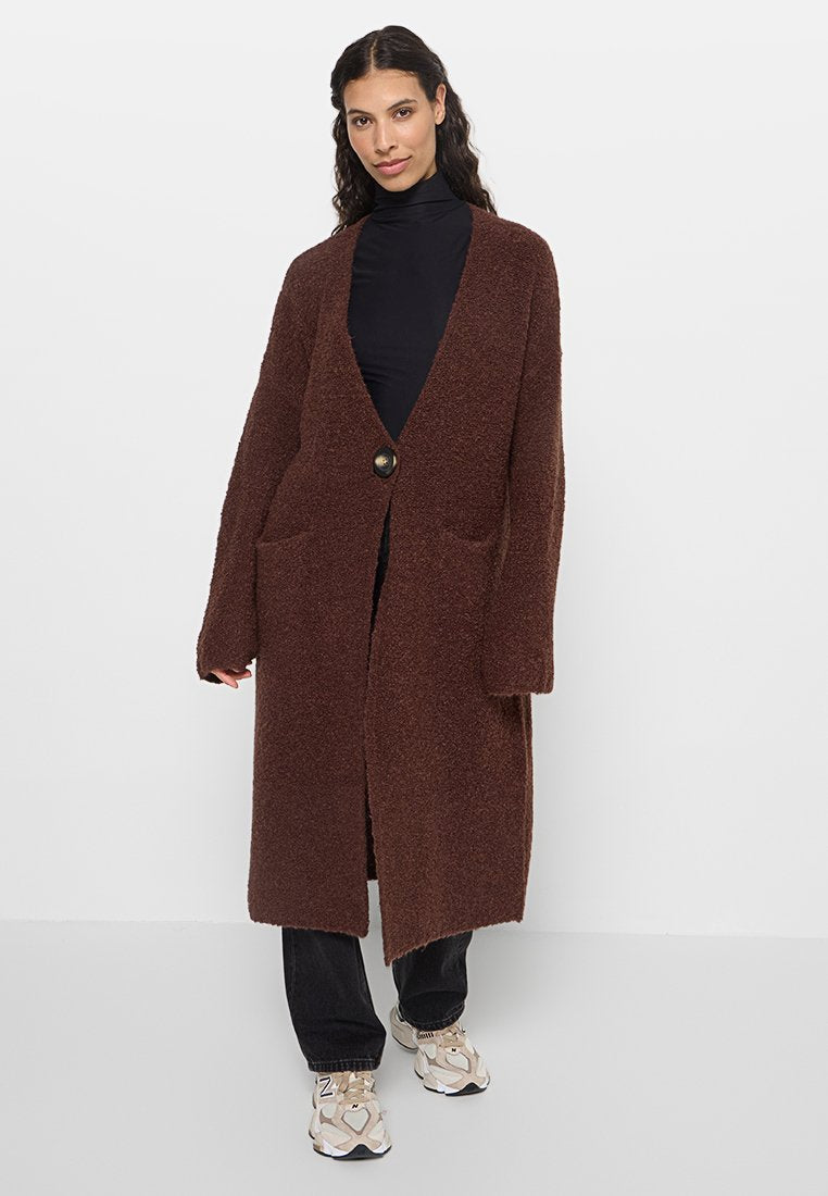 Brauner, flauschiger, langer Cardigan mit tiefem V-Ausschnitt, einzelner Knopfverschluss und Fronttaschen, kombiniert mit einem schwarzen Rollkragenpullover und Sneakers.