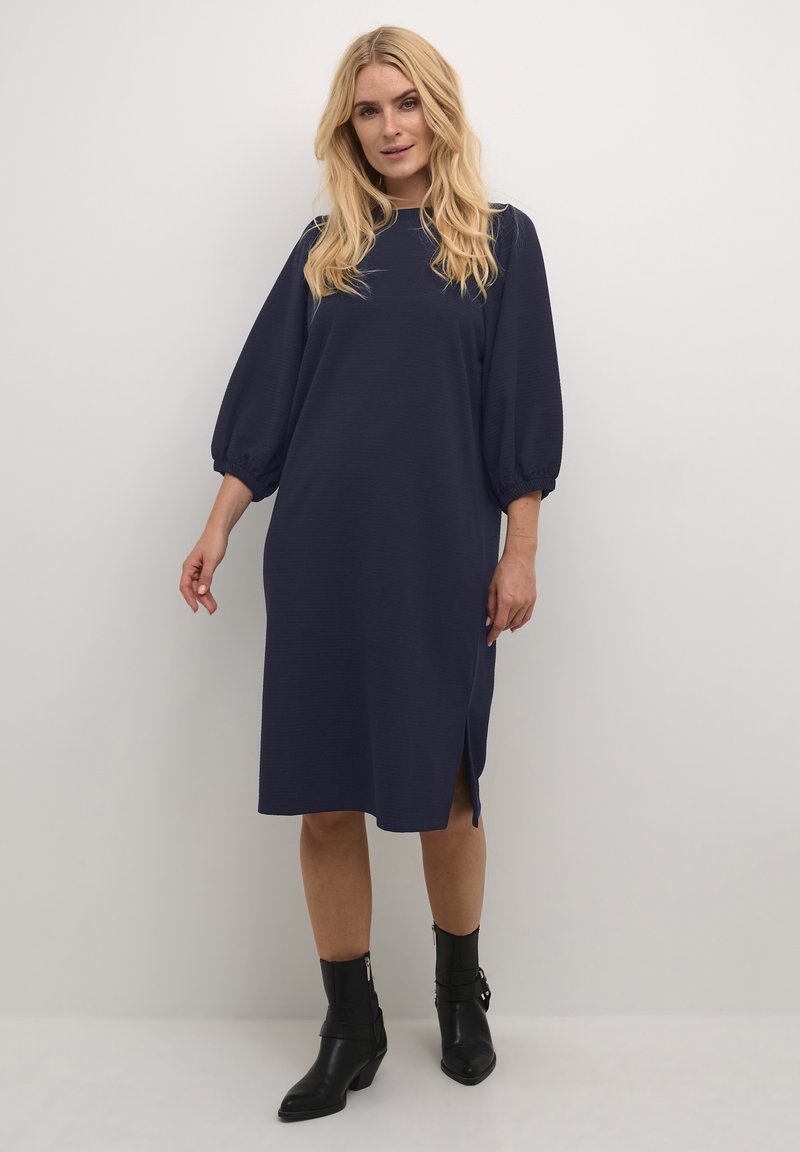 Marineblaues knielanges Kleid mit Puffärmeln, strukturiertem Stoff, seitlichem Schlitz und lockerer Passform. Getragen mit schwarzen Knöchelstiefeln.