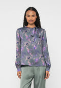 Leichte, transparente Bluse in Grau mit lila Blumenmuster; ausgestattet mit einer Halsbinde und langen Ärmeln mit gesammelten Bündchen. Kombiniert mit grünen Hosen.