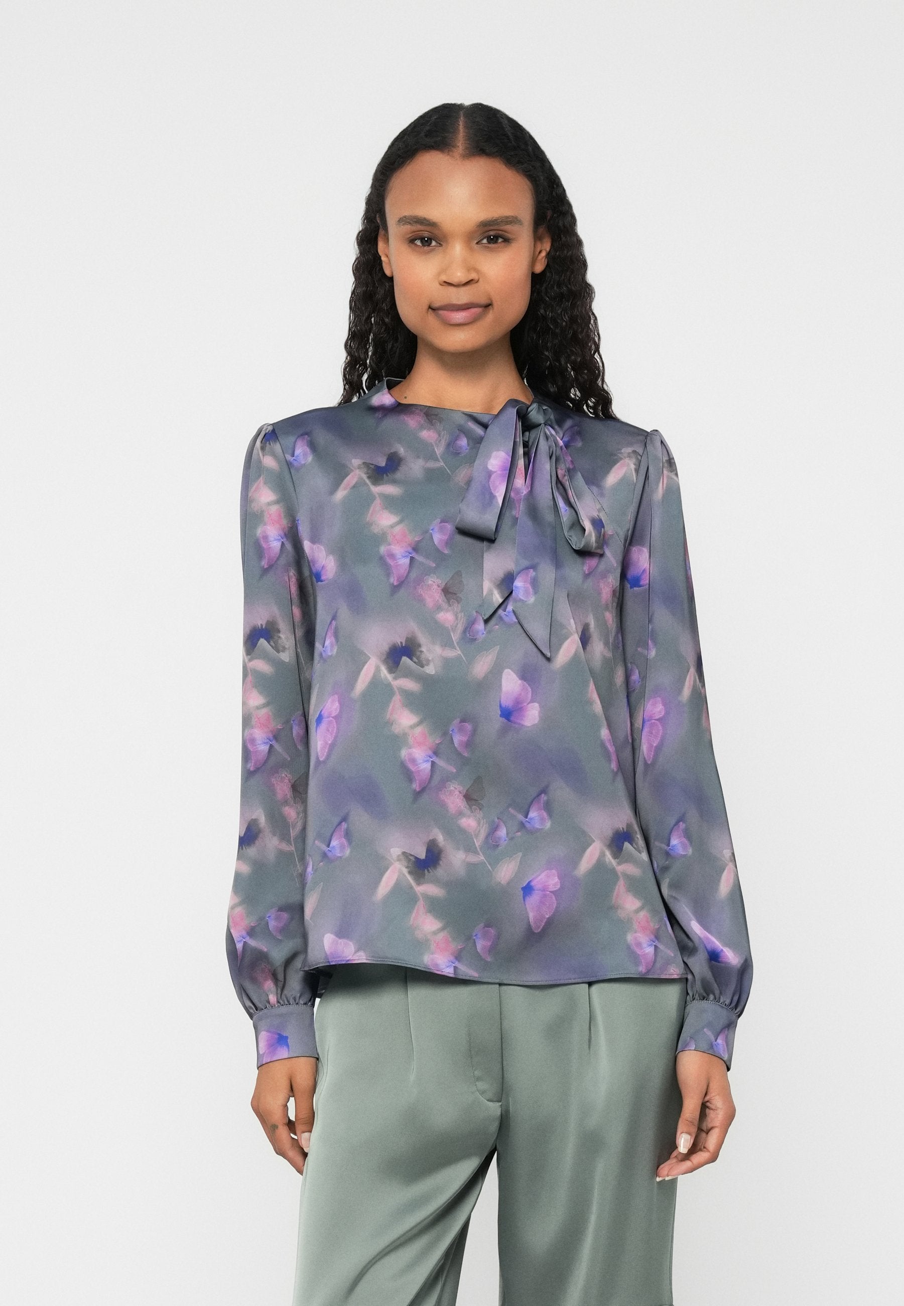 Leichte, transparente Bluse in Grau mit lila Blumenmuster; ausgestattet mit einer Halsbinde und langen Ärmeln mit gesammelten Bündchen. Kombiniert mit grünen Hosen.