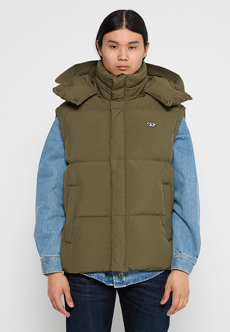 Grüne Steppweste mit hohem Kragen, sichtbaren Nähten und zwei Fronttaschen, getragen über einer blauen Jeansjacke. Mit einem Logo auf der Brust.