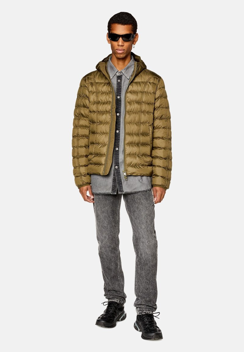 Olivgrüne Steppjacke mit Reißverschluss, getragen über einem grauen Denimhemd, kombiniert mit grauen Jeans und schwarzen Sneakers. Robuste, strukturierte Materialien.