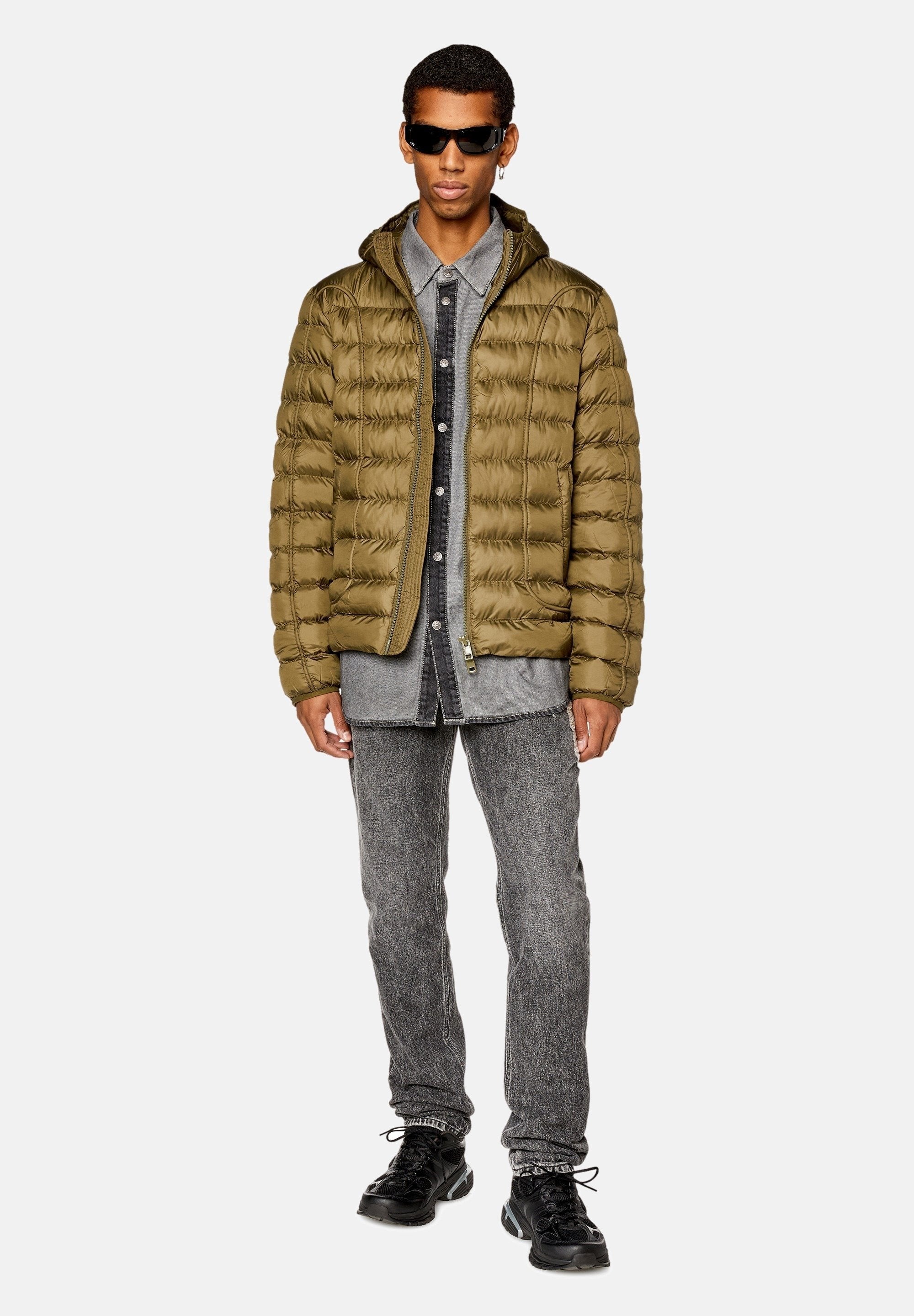 Olivgrüne Steppjacke mit Reißverschluss, getragen über einem grauen Denimhemd, kombiniert mit grauen Jeans und schwarzen Sneakers. Robuste, strukturierte Materialien.