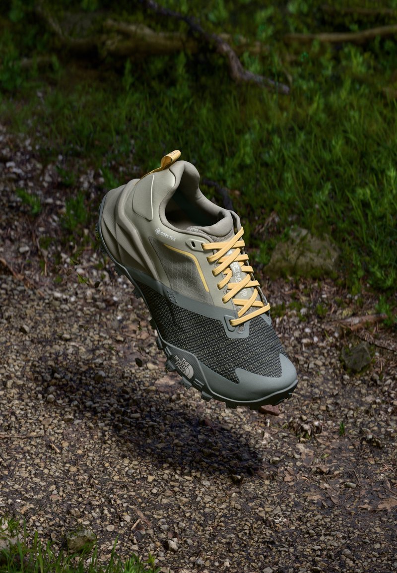 Leichtgewichtiger grauer Gore-Tex-Schuh mit einem strukturierten schwarzen Mesh-Bereich, gelben Schnürsenkeln und gepolsterten Sohlen, erhöht über einer Kiesoberfläche.