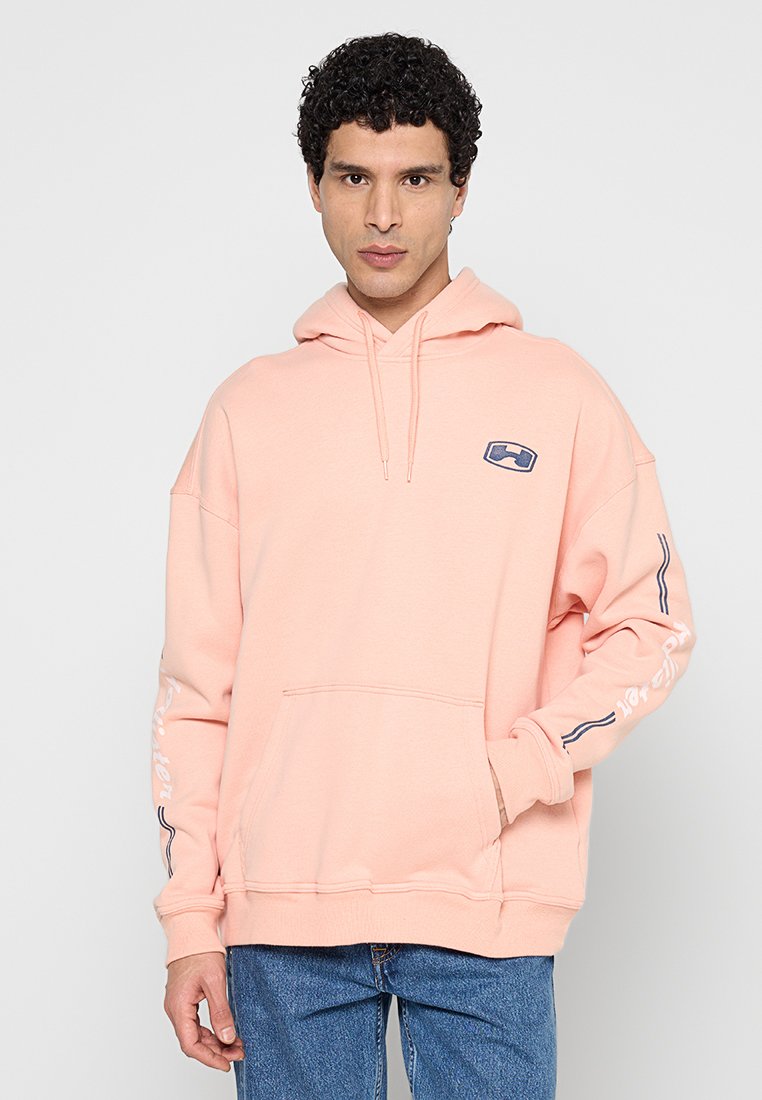 Mann trägt hellrosa Hoodie mit Bauchtasche und blauem Logo auf der Brust, kombiniert mit blauer Jeans, steht vor einfarbigem Hintergrund.