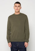 Junger Mann in einem olivgrünen Strickpullover mit kleinem Logo auf der Brust und dunkelblauen Jeans, stehend vor einem schlichten hellen Hintergrund.