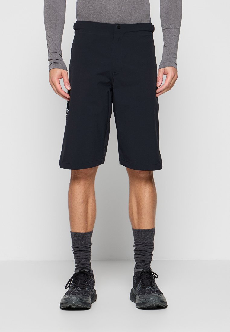 Mann trägt schwarze knielange Shorts, graues Langarmshirt, dunkelgraue Socken und schwarze Sportschuhe und steht vor einem schlichten Hintergrund.