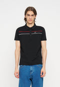 Junger Mann mit schulterlangem braunem Haar, der ein schwarzes kurzärmeliges Polohemd mit roten und weißen Streifen und blaue Jeans vor einem weißen Hintergrund trägt.