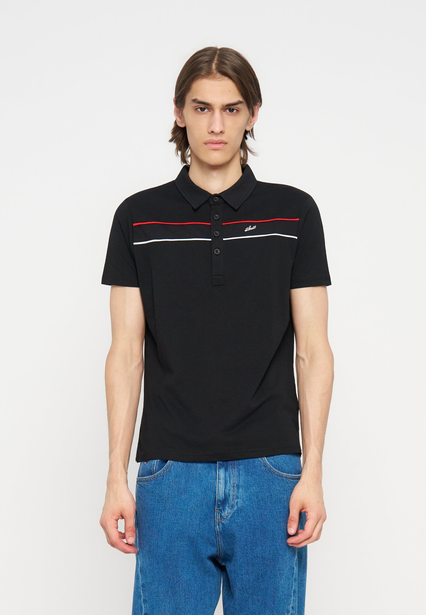 Junger Mann mit schulterlangem braunem Haar, der ein schwarzes kurzärmeliges Polohemd mit roten und weißen Streifen und blaue Jeans vor einem weißen Hintergrund trägt.