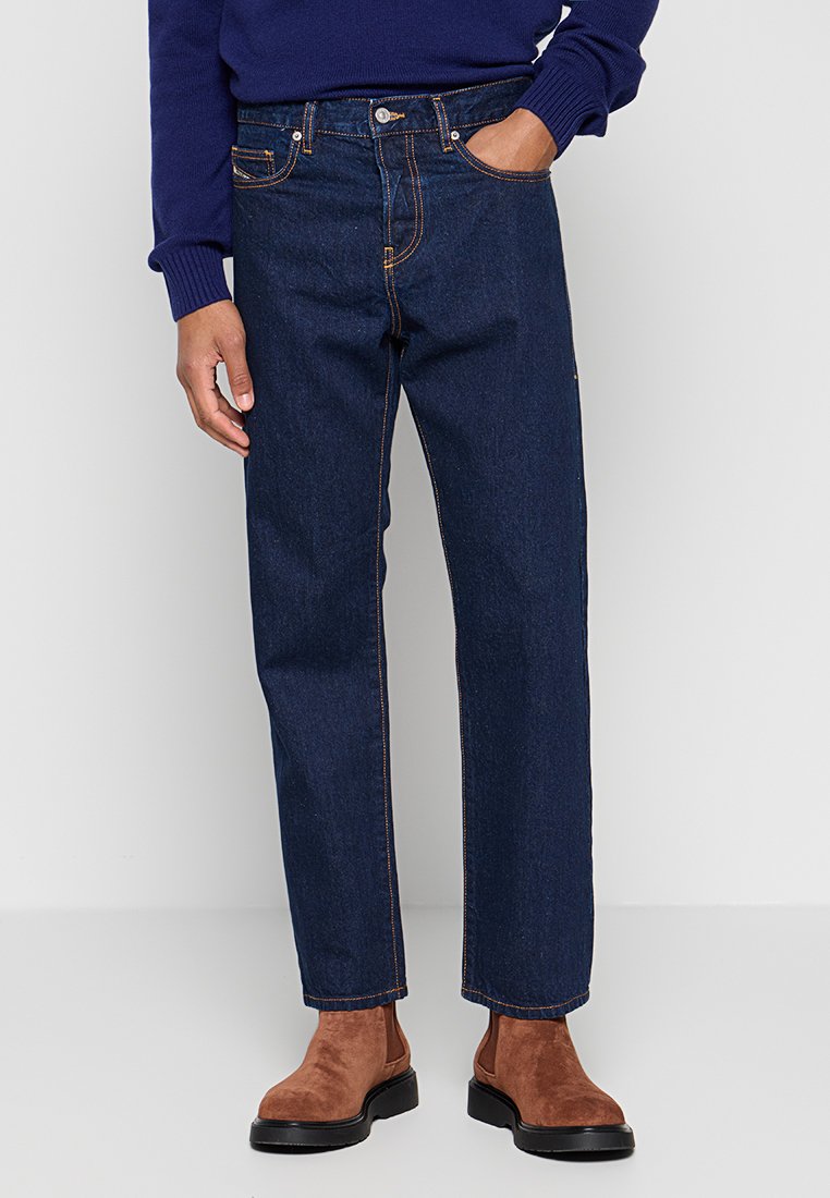 Dunkelblaue Jeans mit geradem Schnitt, mit orangen Nähten, fünf Taschen und einem Knopfverschluss. Getragen mit braunen Wildleder-Stiefeletten.