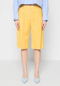 Gelbe, maßgeschneiderte Shorts mit Falten, knielang, aus einem glatten Stoff. Kombiniert mit einem hellblauen Hemd und blauen spitz zulaufenden Absätzen.