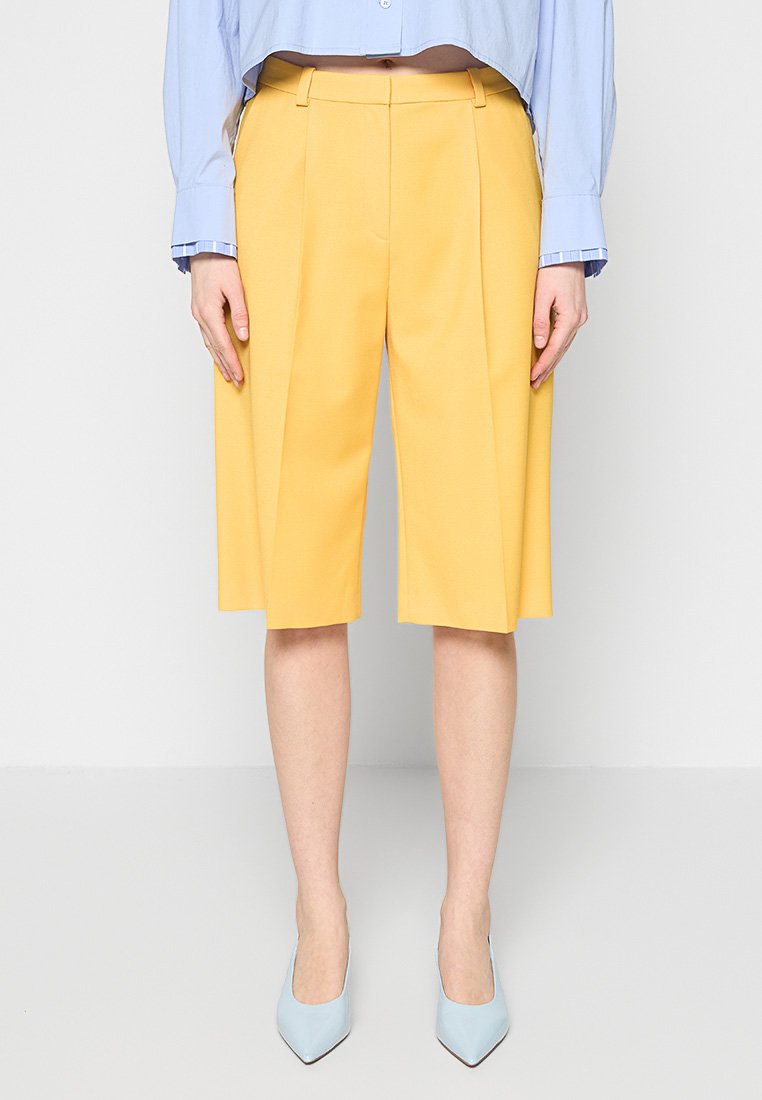 Gelbe, maßgeschneiderte Shorts mit Falten, knielang, aus einem glatten Stoff. Kombiniert mit einem hellblauen Hemd und blauen spitz zulaufenden Absätzen.