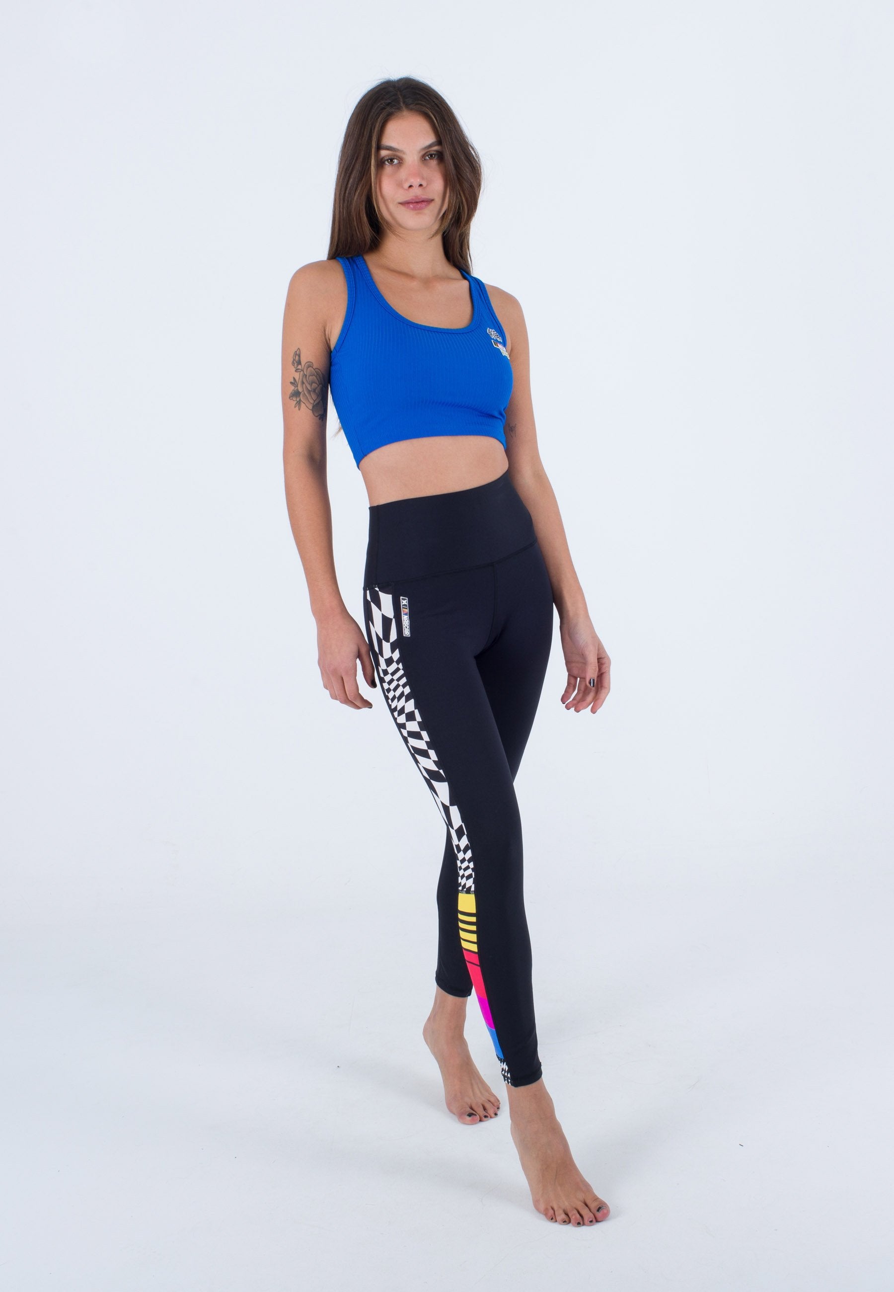 Blaue gerippte Cropped-Bluse kombiniert mit schwarzen Leggings, die ein Schachbrettmuster und farbige Akzentstreifen an den Seiten aufweisen.