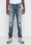 Mann trägt hellblaue, stark abgetragene Slim-Fit-Jeans mit aufgerissenen Knien, ein weißes T-Shirt und weiße Sportschuhe, steht vor einem weißen Hintergrund.