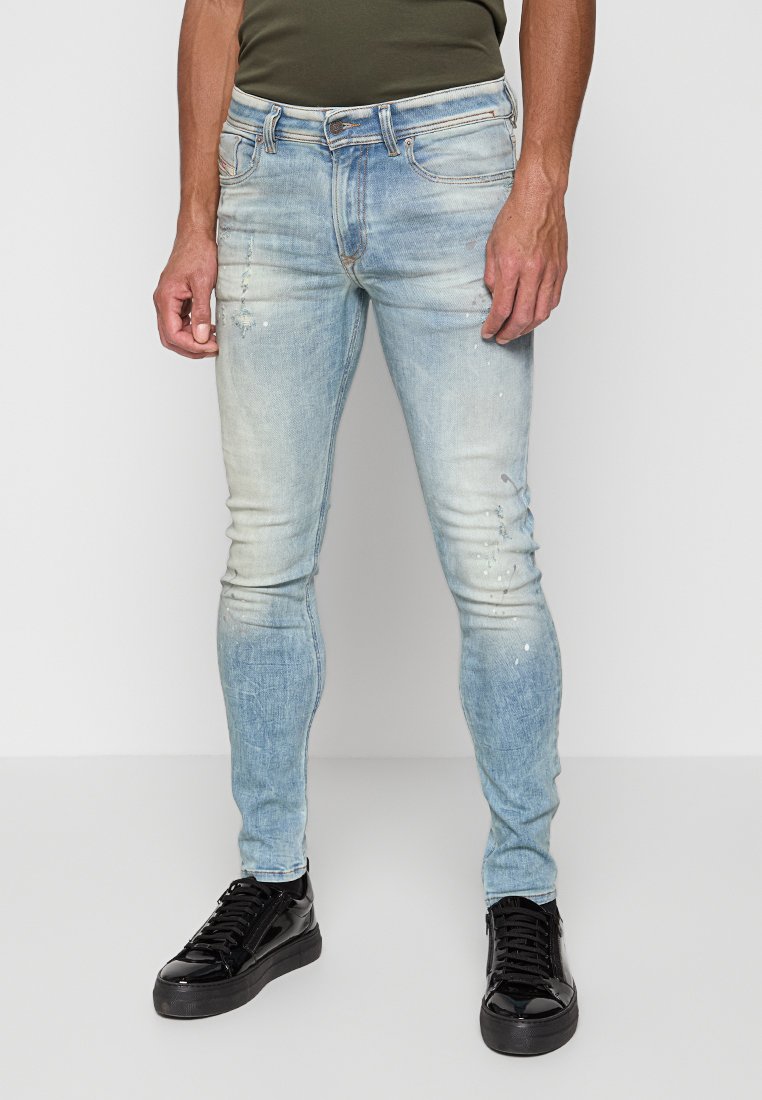 Faded blaue Skinny-Jeans aus Denim, mit abgenutzten Details und Farbspritzern, kombiniert mit schwarzen Turnschuhen.
