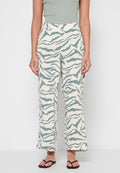 Frau trägt weiße, weit geschnittene Hose mit grünen Zebrastripes, grünes ärmelloses Oberteil und braune Flip-Flops, steht vor einer weißen Wand.