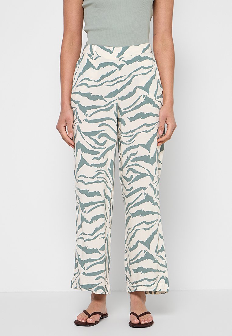 Frau trägt weiße, weit geschnittene Hose mit grünen Zebrastripes, grünes ärmelloses Oberteil und braune Flip-Flops, steht vor einer weißen Wand.