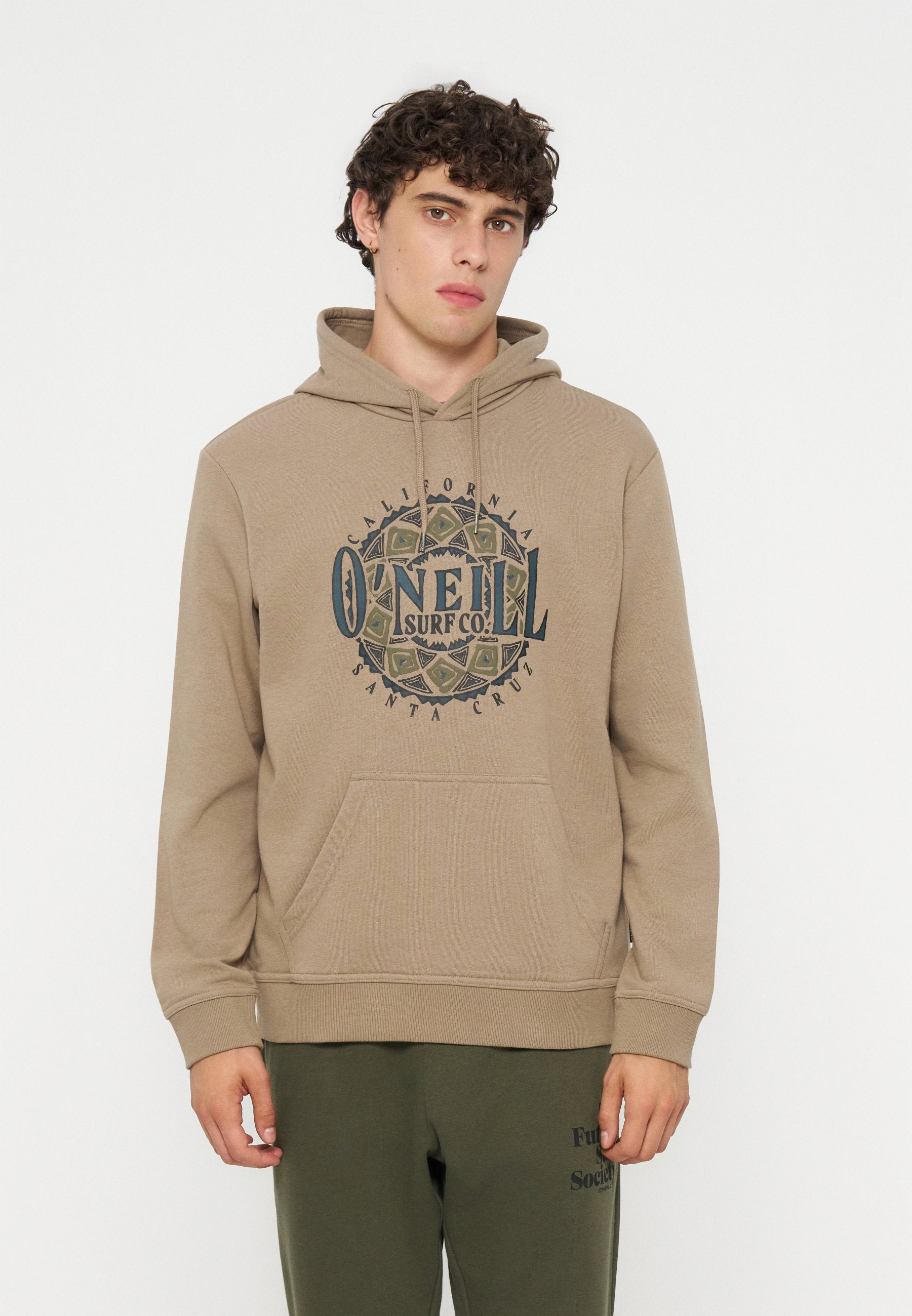Junger Mann mit dunklen lockigen Haaren, der einen beigen O'Neill-Hoodie mit grafischem Text und olivgrüne Hosen vor einem einfarbigen hellen Hintergrund trägt.