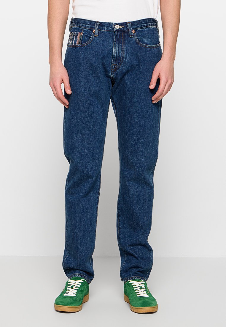 Dunkelblaue Jeans aus Denim mit geradem Bein, fünf Taschen und kontrastierenden Nähten. Kombiniert mit grünen Turnschuhen mit weißen Schnürsenkeln.