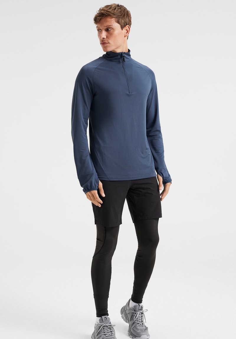 Mann trägt einen navyblauen Langarm-Reißverschlussoberteil, schwarze Shorts über schwarzen Leggings und graue Laufschuhe, steht vor einem weißen Hintergrund.